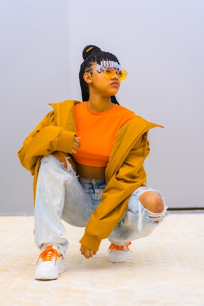 A young trap dancer with braids. black ethnic girl with t-shirt, orange sunglasses and a jacket, crouched looking at camera with seductive face