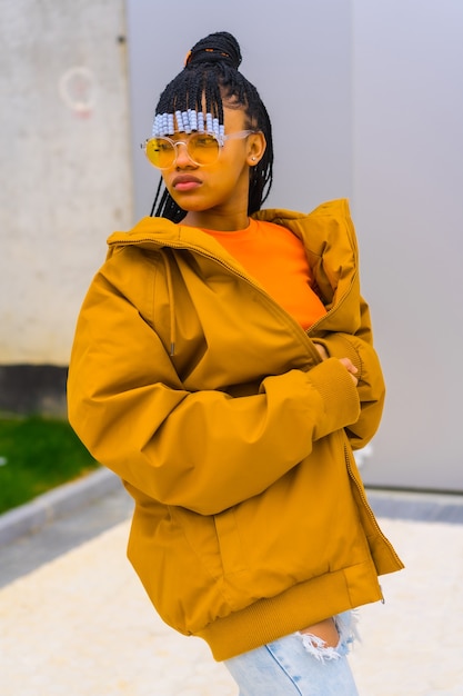 A young trap dancer with braids. Black ethnic girl with t-shirt, orange sunglasses and a jacket, urban post looking at the left