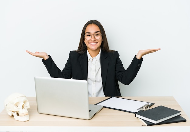 Young traumatologist asian woman isolated on white wall makes scale with arms, feels happy and confident.