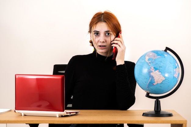 Young travel agent woman sitting behind working desk with laptop computer and geographic globe of the world talking on a cell phone.