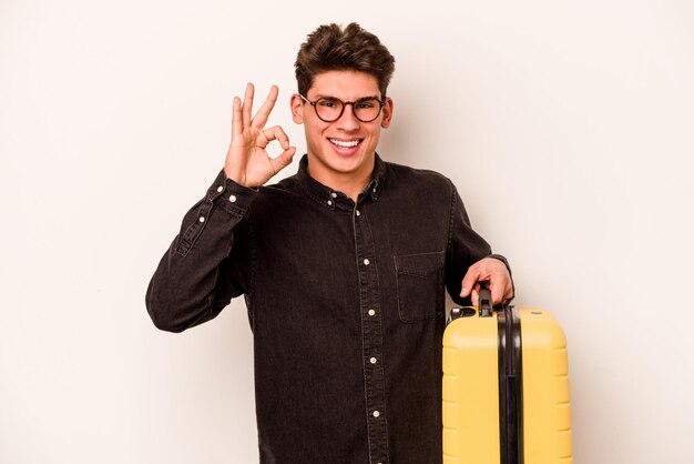 Young traveler caucasian man holding a suitcase isolated on white background cheerful and confident showing ok gesture