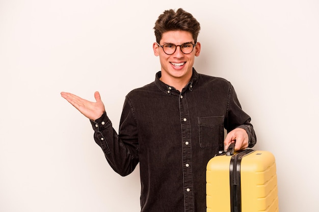 Young traveler caucasian man holding a suitcase isolated on white background showing a copy space on a palm and holding another hand on waist