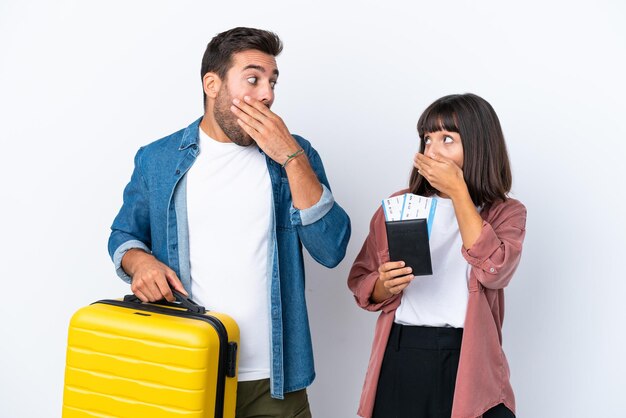 흰색 배경에 격리된 여행가방과 여권을 들고 있는 젊은 여행자 부부는 부적절한 말을 하기 위해 손으로 입을 가리고 있다