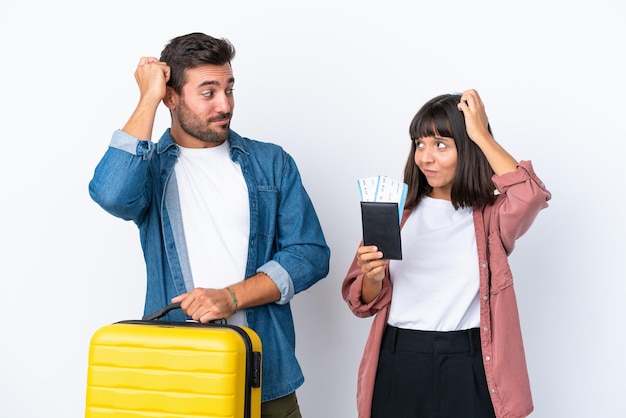 Young traveler couple holding a suitcase and passport isolated on white background having doubts while scratching head