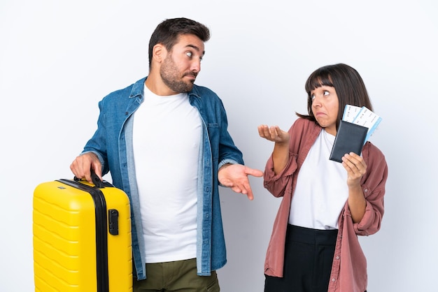흰색 배경에 격리된 여행가방과 여권을 들고 있는 젊은 여행자 부부는 어깨를 들어올리는 동안 중요하지 않은 제스처를 취합니다.