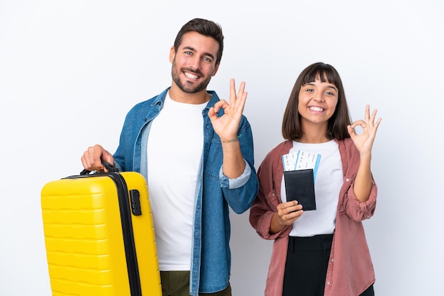 Young traveler couple holding a suitcase and passport isolated on white background showing an ok sign with fingers