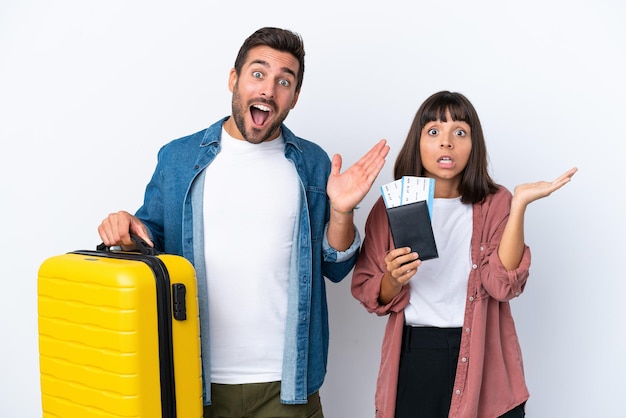 Young traveler couple holding a suitcase and passport isolated on white background with surprise and shocked facial expression