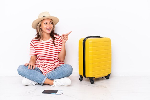 Young traveler girl sitting one the floor isolated on white background pointing finger to the side and presenting a product