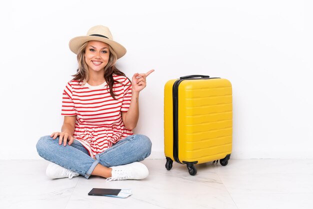 Young traveler girl sitting one the floor isolated on white background pointing finger to the side