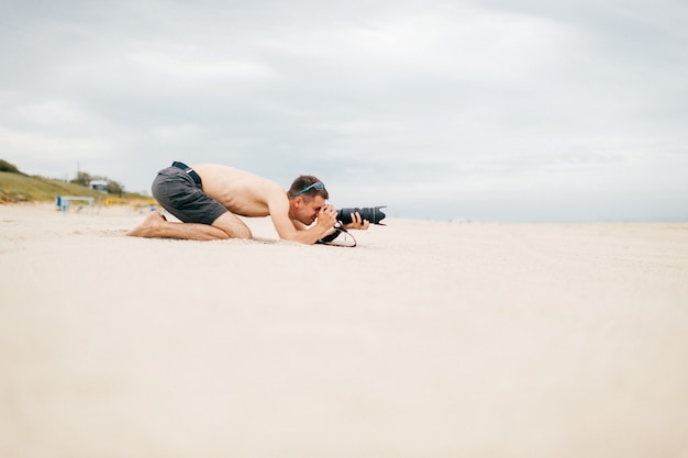 Young traveler man lying on beach near sea on vacation and making photos of nature. Topless handsome barefoot man with camera and big lense spying people from afar. Time for hobby. Boy resting on sand