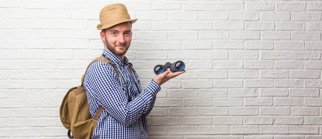 Lo zaino da portare dell'uomo giovane del viaggiatore e una macchina fotografica d'annata che invita per venire, sicuro e sorridente che fa un gesto con la mano, essendo positivo ed amichevole. tenendo un binocolo.