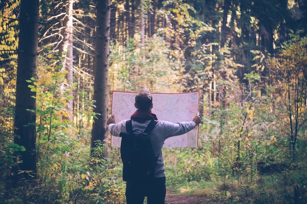 地図、晴れた日に秋の緑の森でバックパックを持つ若い旅行者男。