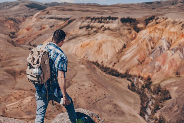 A young traveler in a plaid shirt and with a backpack holds a thermos in his hand and rests his foot on a cobblestone looking at the vast landscape of the canyon.