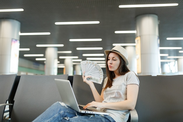 Young traveler tourist woman in hat working on laptop, holding bundle of dollars, cash money while waiting in lobby hall at airport