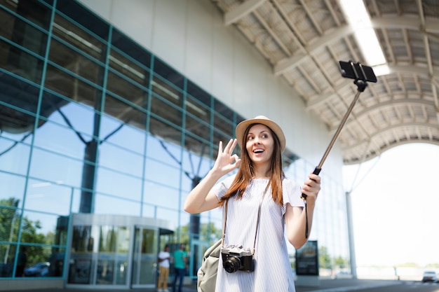 レトロなビンテージ写真カメラを持つ若い旅行者の観光客の女性は空港で一脚の利己的な棒で携帯電話で自分撮りをしているOKサインを表示