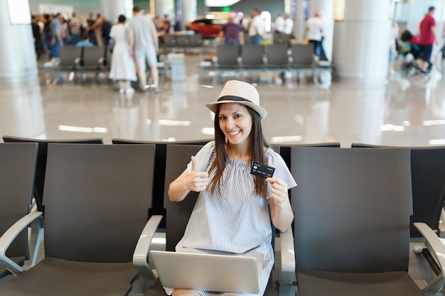 Young traveler tourist woman working on laptop, hold credit card, showing thumb up, waiting in lobby hall at international airport