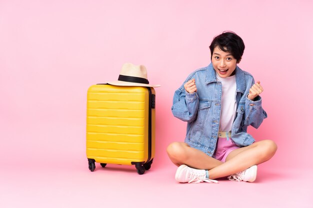 Young traveler Vietnamese woman with suitcase sitting on the floor over pink wall celebrating a victory in winner position