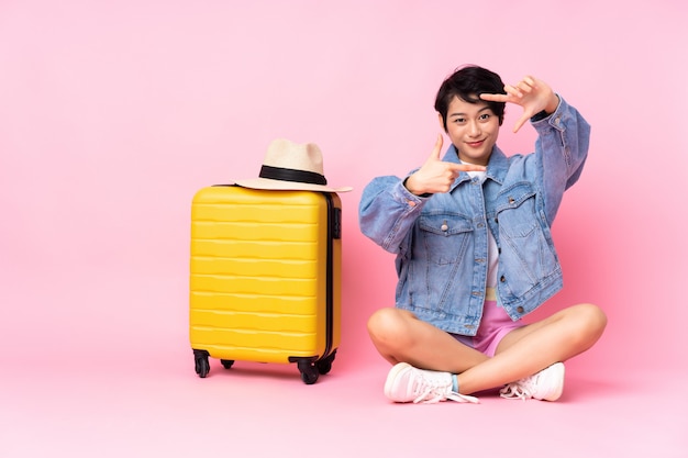 Young traveler Vietnamese woman with suitcase sitting on the floor over pink wall focusing face