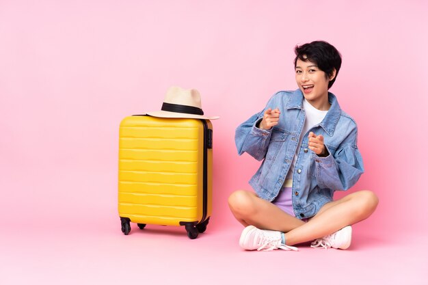 Young traveler Vietnamese woman with suitcase sitting on the floor over pink wall pointing to the front and smiling