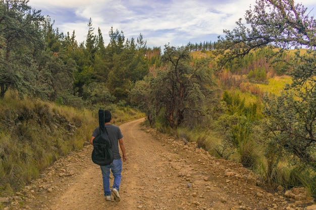 young traveler walking along a mountain trail surrounded by vegetation carrying a guitar on his back