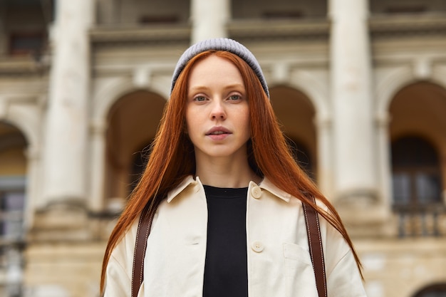 Young traveler with backpack on her shoulder exploring architectural monument place during longawaited summer or autumn vacation wearing coat and hat at cold weather look at camera portrait