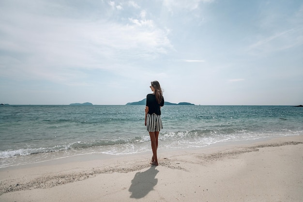 青い海のそばに立って地平線を探している縞模様のスカートと緑のTシャツの若い旅行者の女性。美しい日の出。プーケット。タイ。背面図。