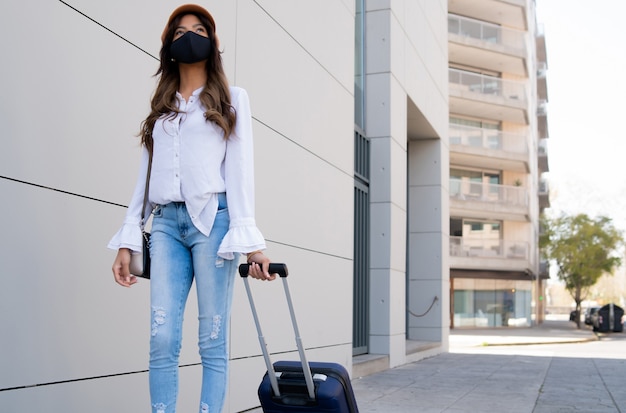 young traveler woman wearing protective mask and carrying suitcase while walking outdoors on the street.