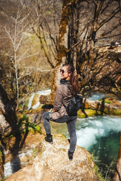 若い旅行中の女性は、マウンテンクリークと美しい滝の上の山の崖の上に立っています。