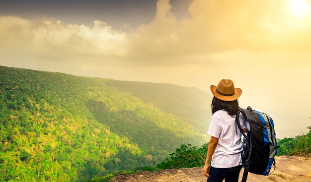 Young traveling woman with backpack and hat stand on the top of the mountain 