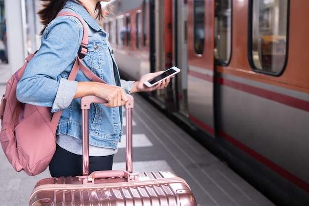 Young traveller girl in jeans jacket with smart phone, pink bag and luggage waiting for the train on the platform, copy space, travel or transportation concept