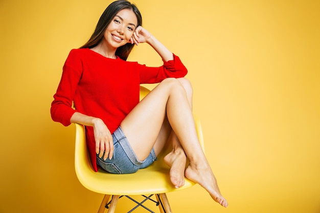 Young trendy beautiful smiling asian woman in red is sitting on chair and possing isolated on yellow background