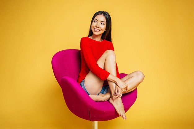 Young trendy beautiful smiling asian woman in red is sitting on chair and possing isolated on yellow background