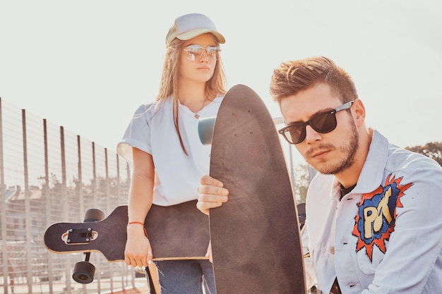 Молодая модная пара отдыхает на солнечной улице со своими лонгбордами. Они носят джинсы.