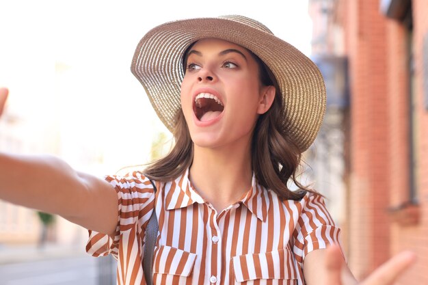 Foto la giovane ragazza alla moda fa il selfie dalle mani con il telefono sulla via della città di estate.