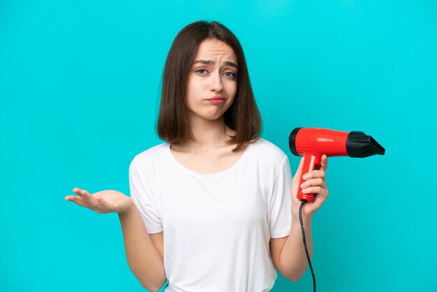 Young Ukrainian holding a hairdryer isolated on blue background making doubts gesture while lifting the shoulders