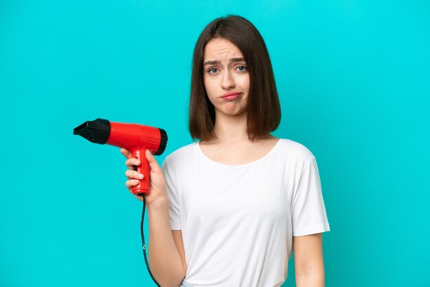 Young Ukrainian holding a hairdryer isolated on blue background with sad expression