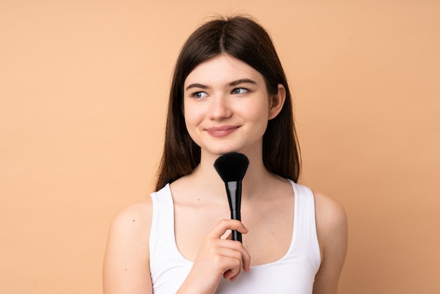 Young Ukrainian teenager girl over isolated wall holding makeup brush and thinking