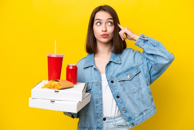 Young Ukrainian woman holding fast food isolated on yellow background having doubts and thinking