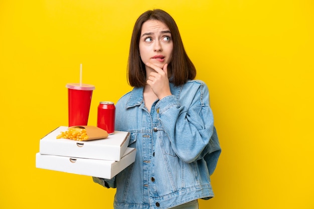 Foto giovane donna ucraina che tiene un fast food isolato su uno sfondo giallo con dubbi e pensieri