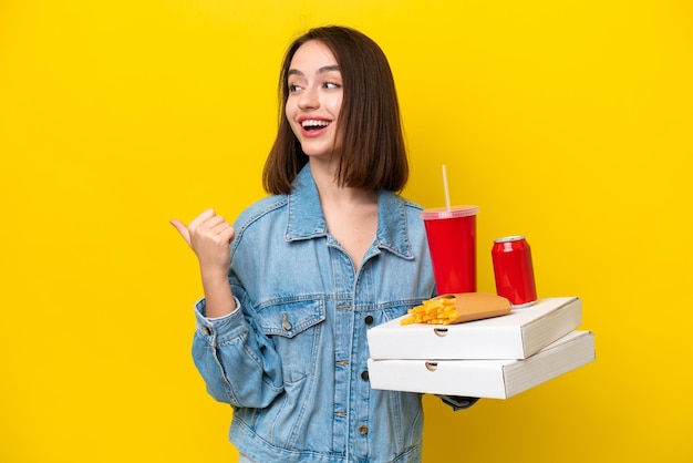 Young Ukrainian woman holding fast food isolated on yellow background pointing to the side to present a product