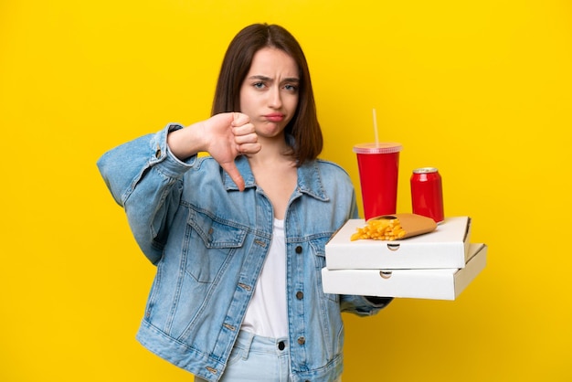 Young ukrainian woman holding fast food isolated on yellow background showing thumb down with negative expression