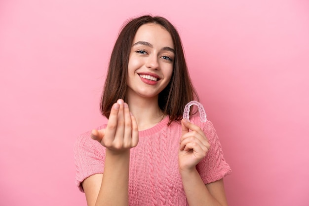 Young Ukrainian woman holding invisible braces isolated on pink background inviting to come with hand Happy that you came