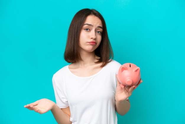 Young ukrainian woman holding a piggybank isolated on blue background making doubts gesture while lifting the shoulders