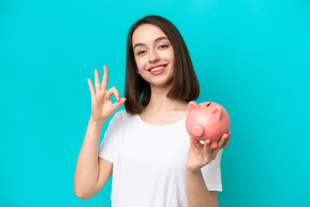 Young Ukrainian woman holding a piggybank isolated on blue background showing ok sign with fingers