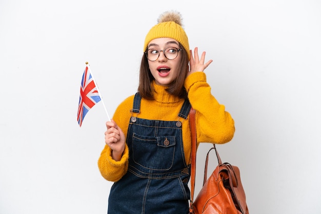 Young Ukrainian woman holding an United Kingdom flag isolated on white background listening to something by putting hand on the ear