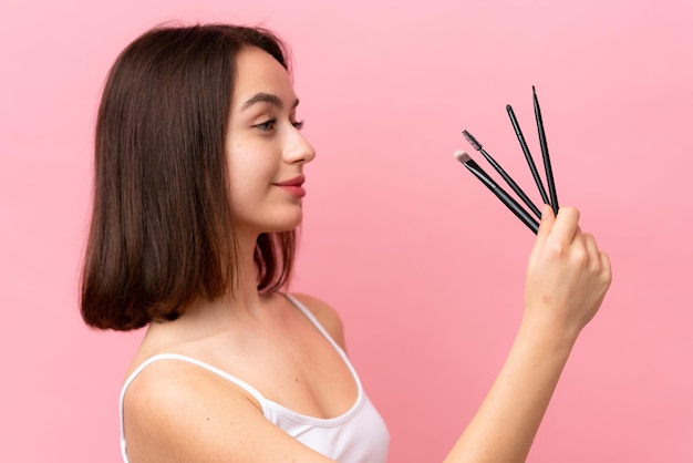 Young Ukrainian woman isolated on pink background holding makeup brush and lookin it