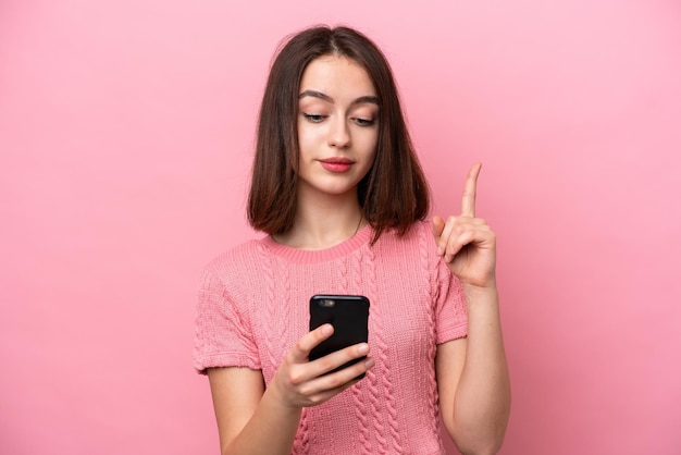 Young Ukrainian woman isolated on pink background using mobile phone and lifting finger