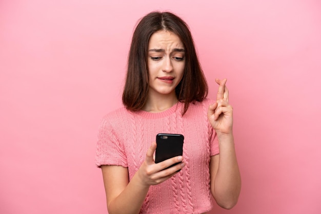 Young ukrainian woman isolated on pink background using mobile\
phone with fingers crossing