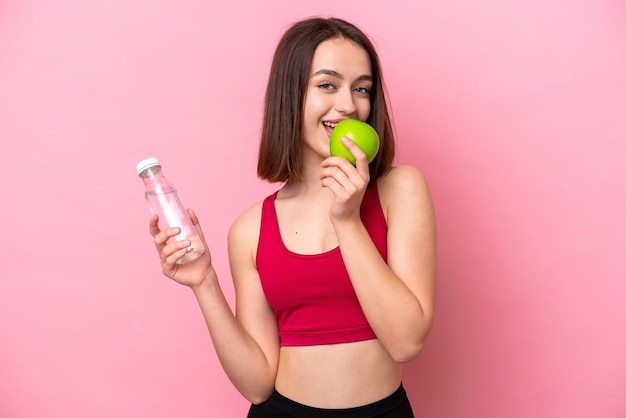 Young Ukrainian woman isolated on pink background with a bottle of water and eating an apple