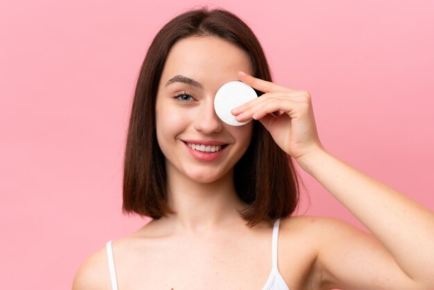 Young Ukrainian woman isolated on pink background with cotton pad for removing makeup from her face and smiling